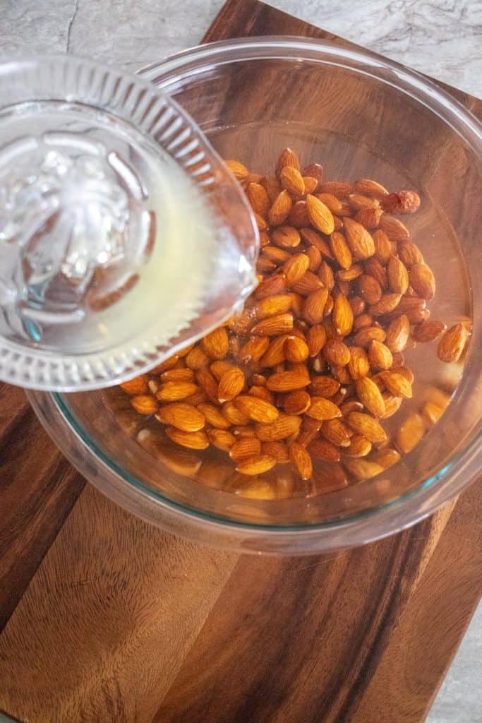 Lemon juice in a glass juicer, being poured over almonds soaking in water