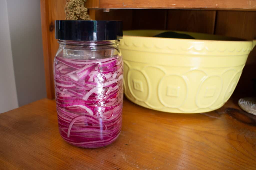 Jar of red onions in brine next to a bright yellow fruit bowl