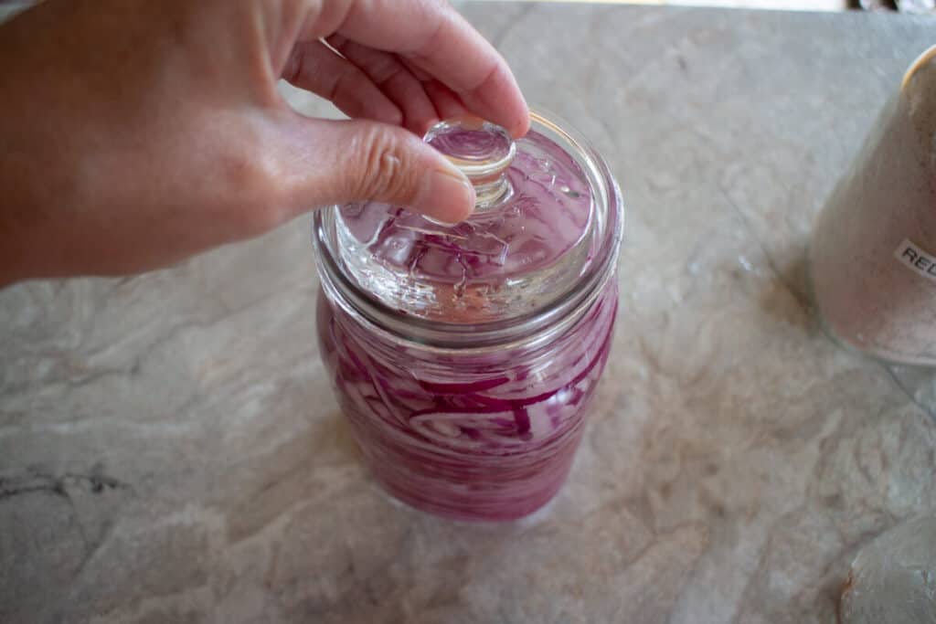 Placing glass fermentation weight on top of red onions in brine