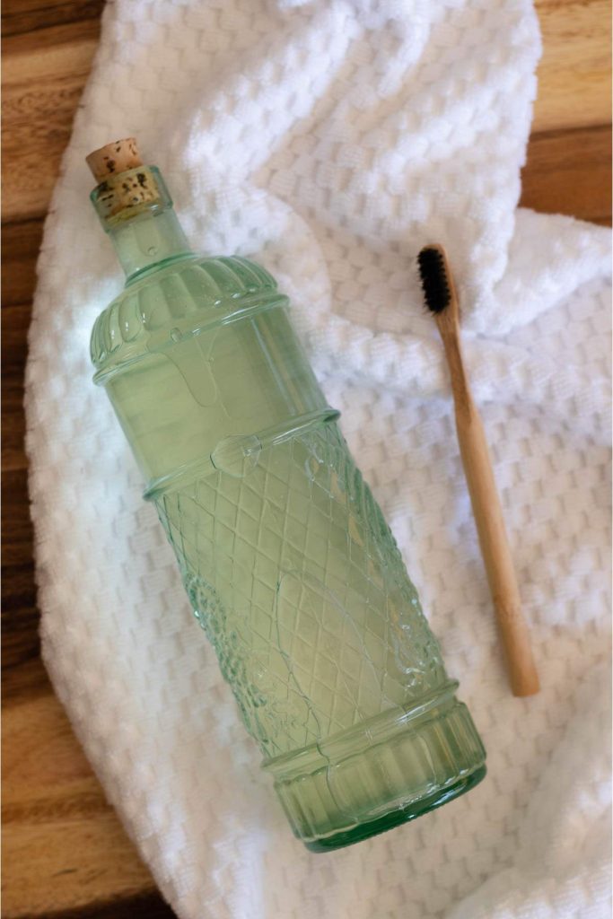 green glass bottle of homemade mouthwash, and a bamboo toothbrush on a white towel