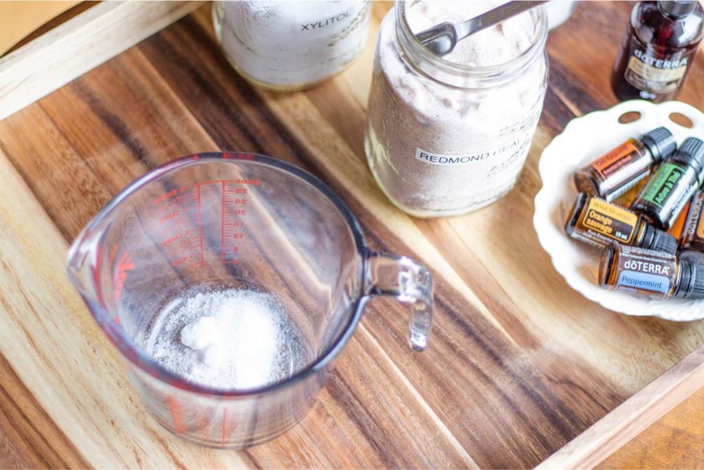 glass measuring cup containing baking soda, xylitol and sea salt, on a wooden tray