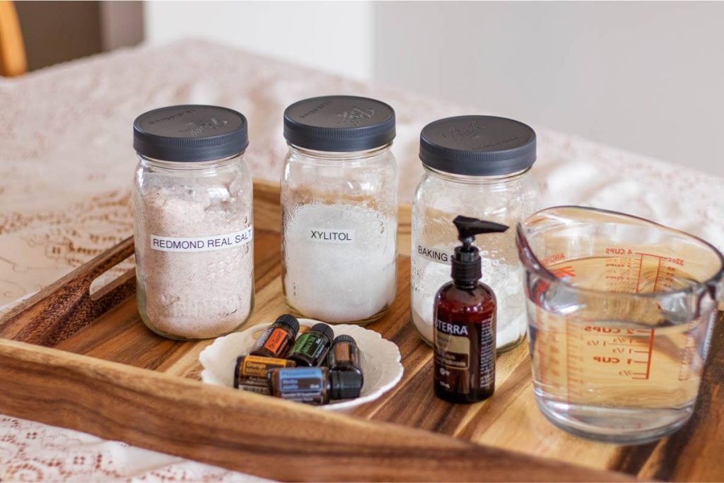 jars of sea salt, baking soda and xylitol, some essential oils, coconut oil and a glass measuring cup on a wooden tray